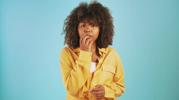 Darkskinned Female Looking Scared and Nervous Touching Her Lips While Posing Against Blue Studio