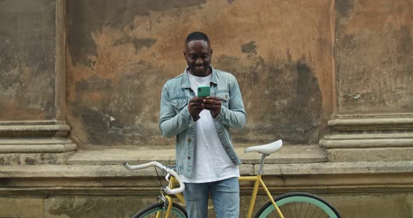 Young Afro American Man Wearing Casual Clothes Using Smartphone, Looking To the Screen and Smiling