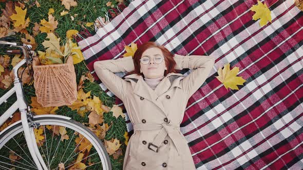 Young Woman with Closed Eyes Smiling Lying on Checkered Rug and Green Grass Covered By Yellow Leaves