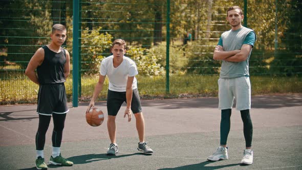 Three Sportsmens Standing on the Basketball Court Outdoors - One Sportsman Hitting the Ball Off the