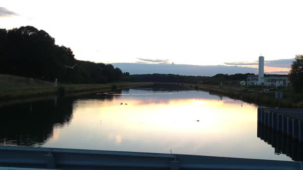 Drone pans over the edge of a damn while birds fly over the reflection of the sunset.