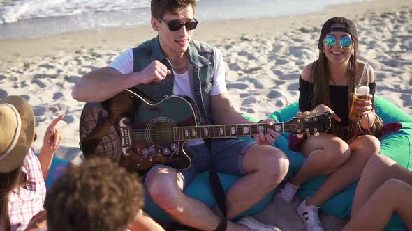 Young Man Playing Guitar Among Group of Friends Sitting on Easychairs on the Beach and Singing on a