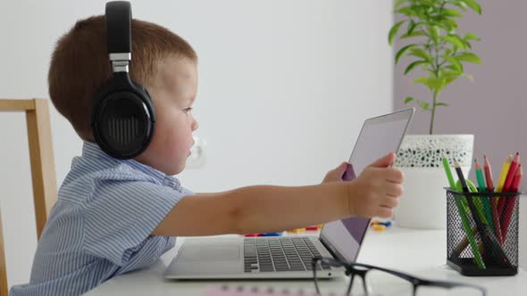 Caucasian Little Elementary Pupil Sitting at Table in Headphones Opening Laptop
