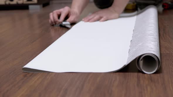 Male Sitting on Floor Cuts Off a Strip of Wallpaper for Pasting with a Knife