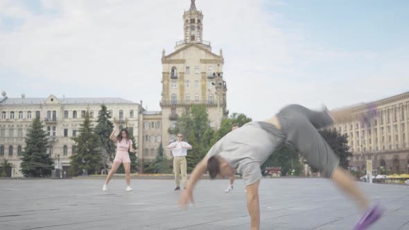 Brunette Young Man Performing Sportive Dancing Movements on City Square with Friends Dancing at the