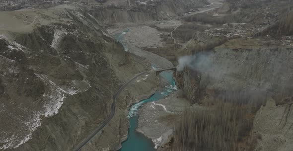 Aerial Over Turquoise Colour River In Hunza Valley With Smoke Seen Rising Near Bridge. Dolly Forward