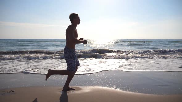 Young Sporty Man Running on the Sea Beach at Morning. Athletic Guy Jogging Along Coast on a Sunny