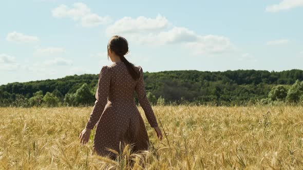 Attractive Young Woman in Brown Dress Walks Along Yellow Field Against Blue Sky with Clouds