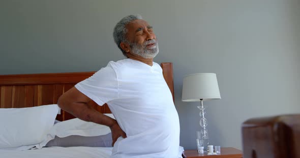 Front view of senior black man sitting on bed holding lower back in a comfortable home 4k