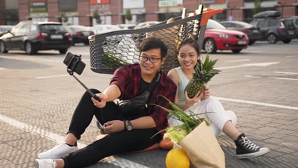 Satisfied Young Asian Couple Sitting in the Ground Near Market Trolley