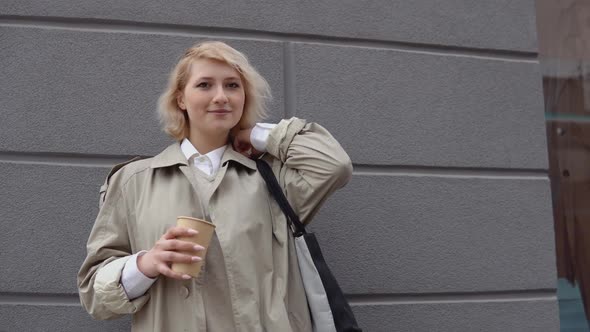 Young Blonde Business Woman in a Beige Trench Coat and White Blouse with a Black Bag and Takeaway