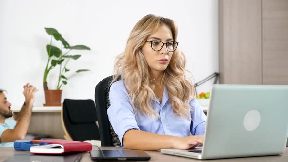 Freelancer Woman Working on the Computer While Her Husband Is Watching a Ball Game in the Background