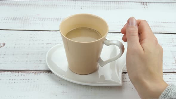 The Girl's Hand Takes a Cup of Coffee Close-up. Coffee Cup on a Saucer in the Shape of a Heart on a