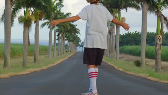 A Teenager with Long Hair Rides a Skateboard Along a Beautiful Road with Palm Trees