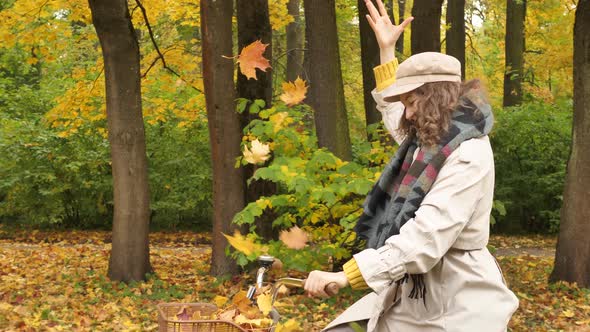 Happy Young Woman Rides a Bicycle with a Basket Through the Autumn Park and Throws Fallen Maple