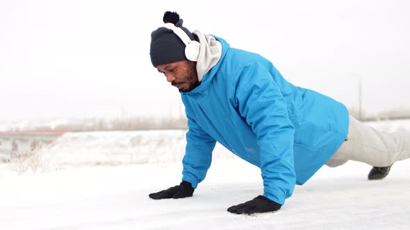 Black Man in Warm Clothes and Headphones Trains Outdoors in Winter Doing Pushups Side View