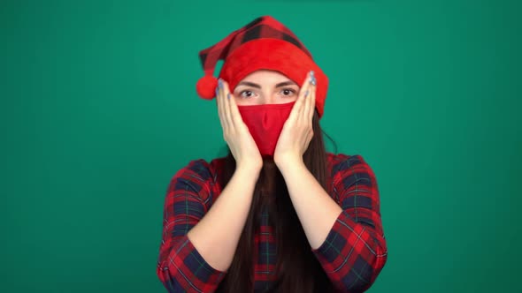 Young Woman in Santa Hat and Red Medical Mask with Shocked Expression Keeps Palms on Cheek