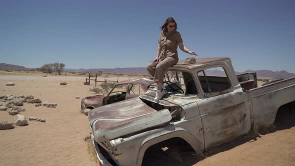 Young Woman in Safari Overalls Sits on the Rooftop of Abandoned Old Rusty Car