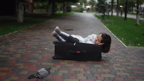 Wide Shot Young Little Woman Sleeping Lying in Suitcase on City Street with Pigeon Walking at Front