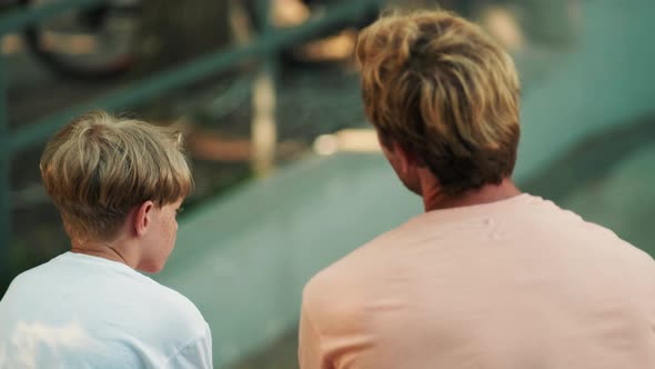 A back view of a father is talking to his son sitting on the sports ground at summer