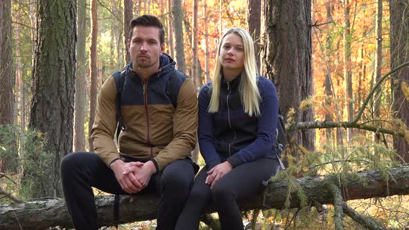 A Hiking Couple Sits on a Broken Tree and Looks Seriously at the Camera in a Forest