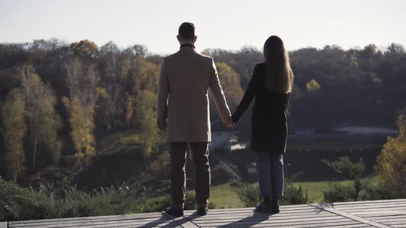 Back View of Caucasian Man and Woman Holding Hands Standing on Wooden Bridge and Looking Away at