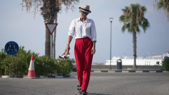 Exhausted Caucasian Woman Walking on Urban Embankment on Hot Summer Day Holding Elegant Highheels in