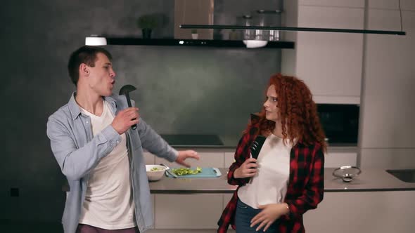 Happy Carefree Young Family Couple Husband and Wife Singing in Soup Ladle and Pasta Tongs Microphone