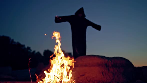 Evil witch in black cloak with hood near the fire. Halloween costume.