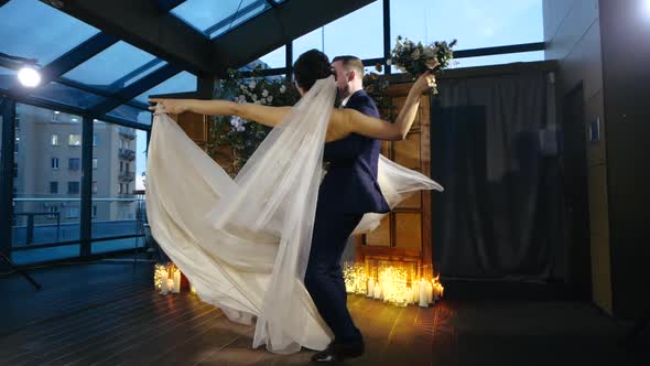 Bearded Groom in Suit Holds His Beloved Bride in Beautiful Dress on Arms and with Bouquet of Flowers