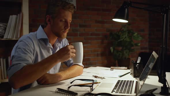 Young Architect with Blue Shirt and Light Beard Drinking From White Cup That Putting on Glasses at