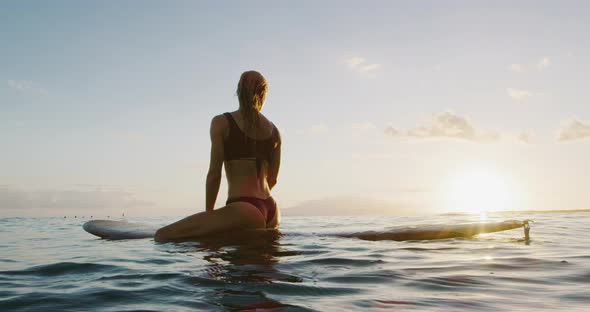 Young woman sitting on a surfboard at sunset alt