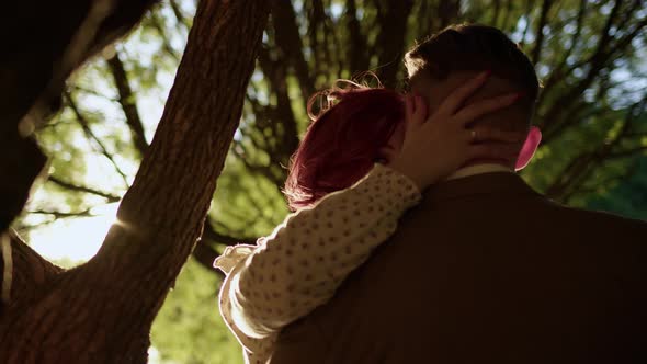 Unrecognizable Couple of Man and Woman Hugging Under Tree and Kissing Against Backdrop of Setting
