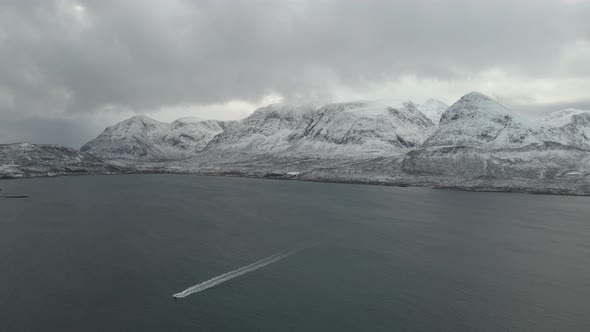 High angle drone view of boat speeding along calm ocean, mountains covered in snow