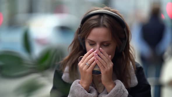 Young Inspired Woman Is Warming By Hot Coffee in Street Cafe, Listening To Music By Headphones