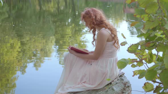 Side View of the Redhead Attractive Caucasian Girl Reading the Book in Dark Red Cover on the Bank of