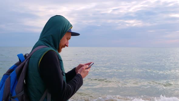 Bearded man in raincoat on a blue sea landscape background with smartphone in hands texting