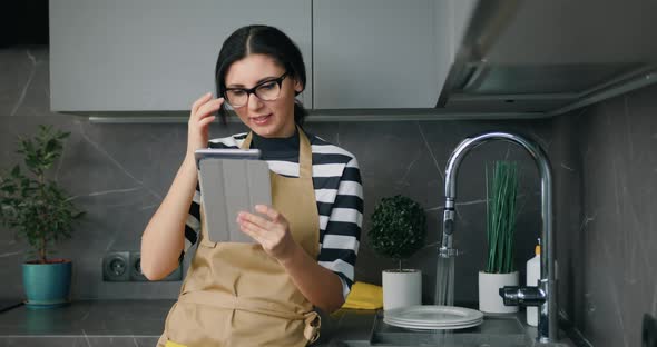 Beautiful Woman in Eyeglasses Using Digital Tablet Screen when Washing Dishes at Kitchen Home