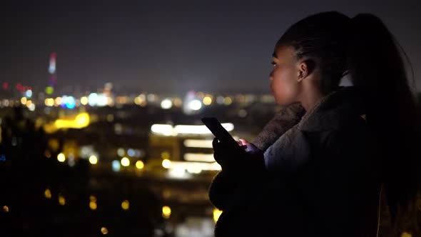 A Young Black Woman Works on a Smartphone and Looks at the Cityscape