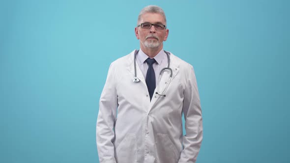 Doctor in Glasses and a Robe with a Stethoscope Shrugs in the Studio a Blue Background