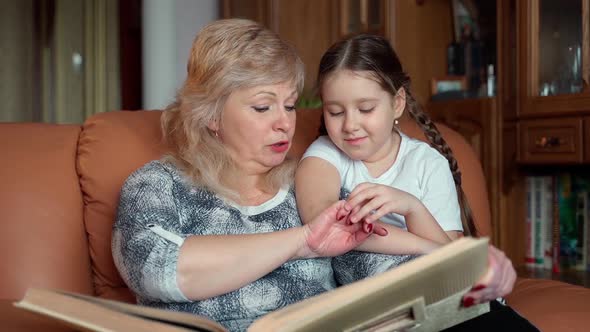 Warm slow motion shot senior woman shows little girl family album sitting at home indoors