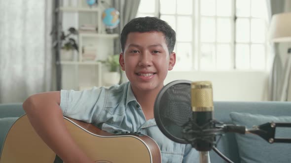 Asian Boy With Guitar And Microphone Smiling. The Child Is Broadcasting Live On The Internet