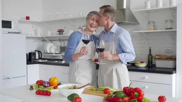 Happy Old Couple Drinking Wine Preparing Vegetable Salad in Kitchen at Home