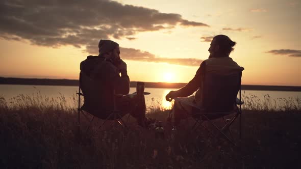 Camping Men Having a Meal Near Water During Sunset Sitting in Chairs