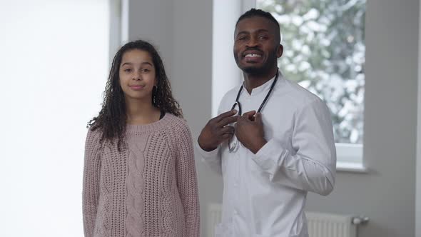 Portrait of Positive African American Doctor and Teen Patient Looking at Camera Smiling Standing