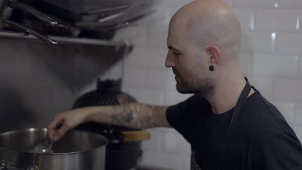 Tattooed Male Chef Tastes Food with Spoon in Kitchen Close Slomo