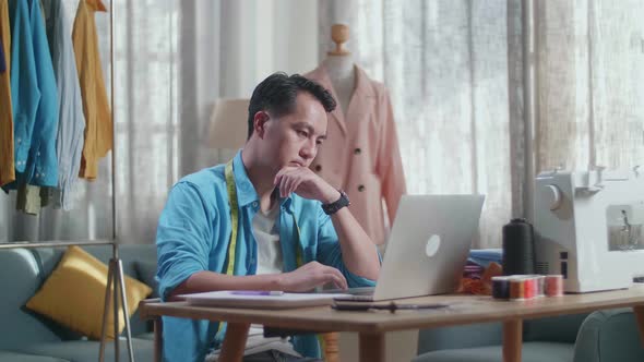 Asian Male Designer Working On A Laptop And Thinking While Designing Clothes In The Studio