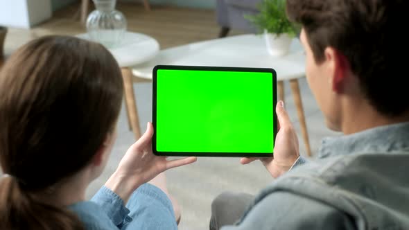 Over The Shoulder View Young Couple at Home Sitting on a Sofa Using With Green Mock-up Screen Tablet