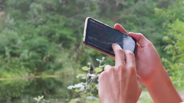 A Male Analyzes the Graph of a Cryptocurrency in a Smartphone in Nature