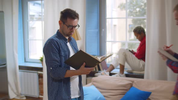 Young Guy Hipster Reading Textbook Walking in Dorm Room with Other Students Study